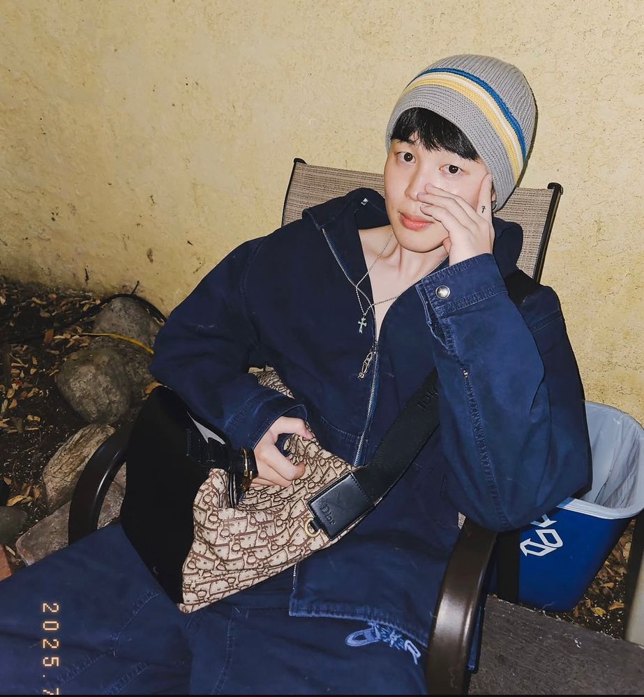 jimin in a denim dior outfit as he sits outside. his fingers cover half his face as he looks into the camera. he has on a gray beanie with a blue, white, and yellow stripe 