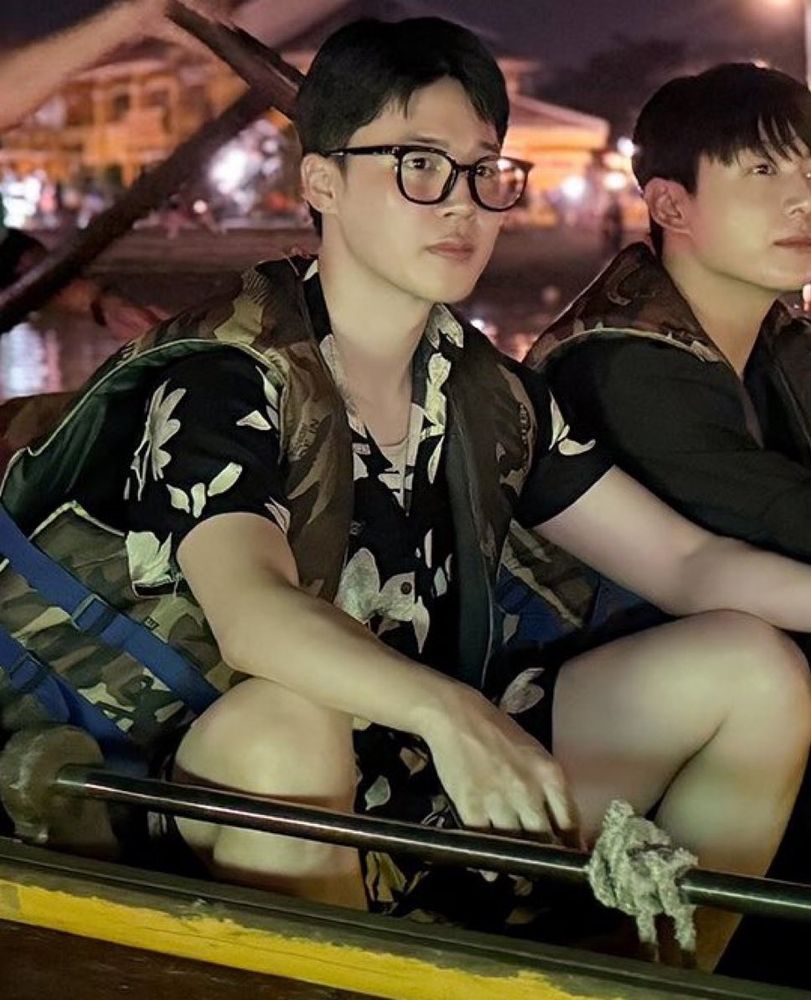 jm with short black hair and glasses as he sits on a boat next to jungkook for ays 2. he has on a black and white floral matching short and shirt set.