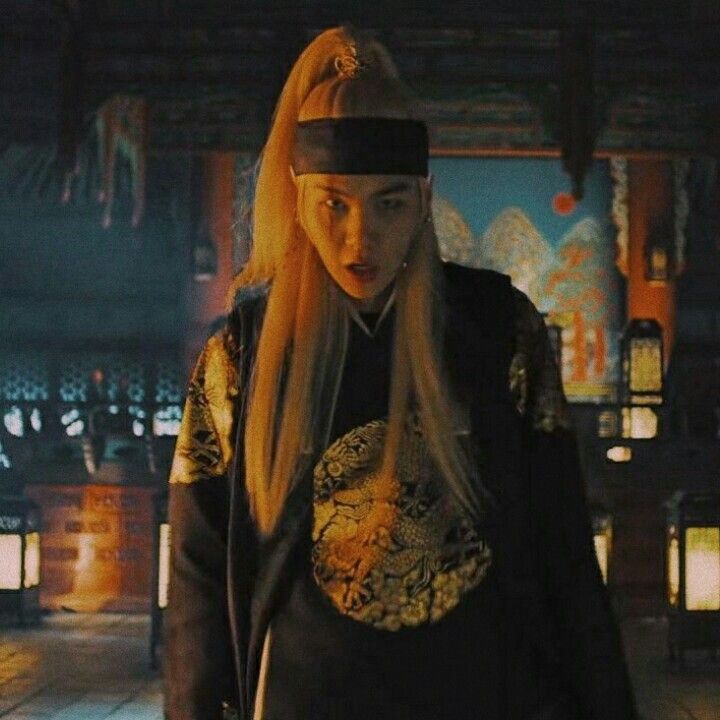 agust d mid frame as he walks close to the camera. he has long blond hair and a headband with a black and gold robe.