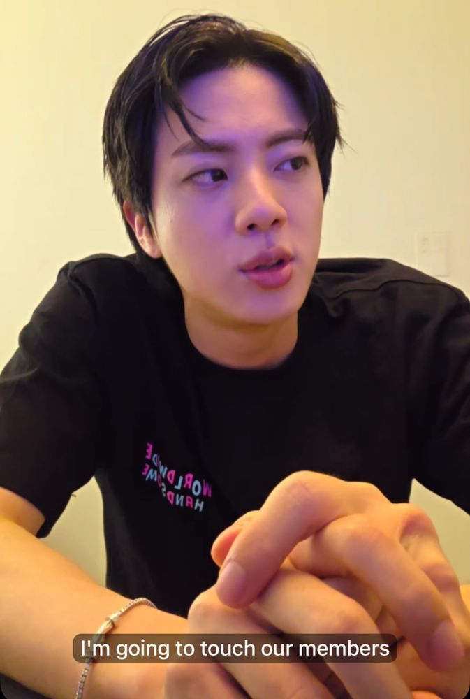 screenshot from jin’s recent live. his hair is parted and he wears a black shirt with his hands on the table. the rough (very rough) translation says; “i’m going to touch our members.” joke on the funny translation 
