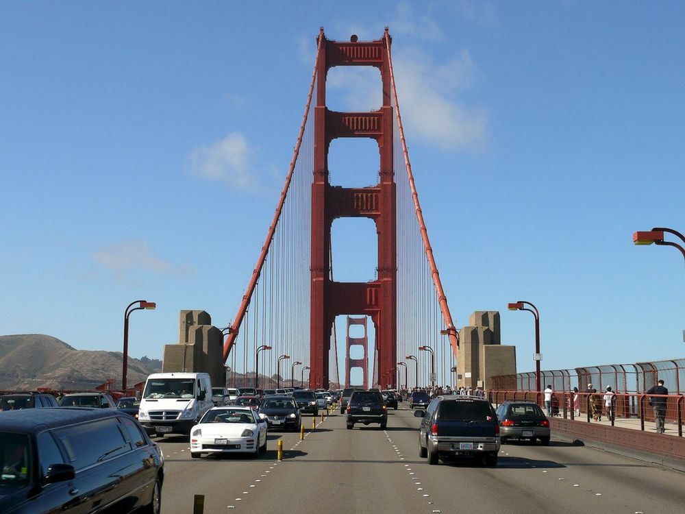 View from the roadway of the Golden Gate Bridge in San Francisco, showing heavy traffic approaching the iconic red-orange suspension bridge tower. The road is divided by yellow movable plastic sticks, and the right side features a pedestrian walkway separated by orange safety barriers. Multiple lanes of cars and trucks travel toward the towering bridge structure under a blue sky with white clouds, with hills visible in the background