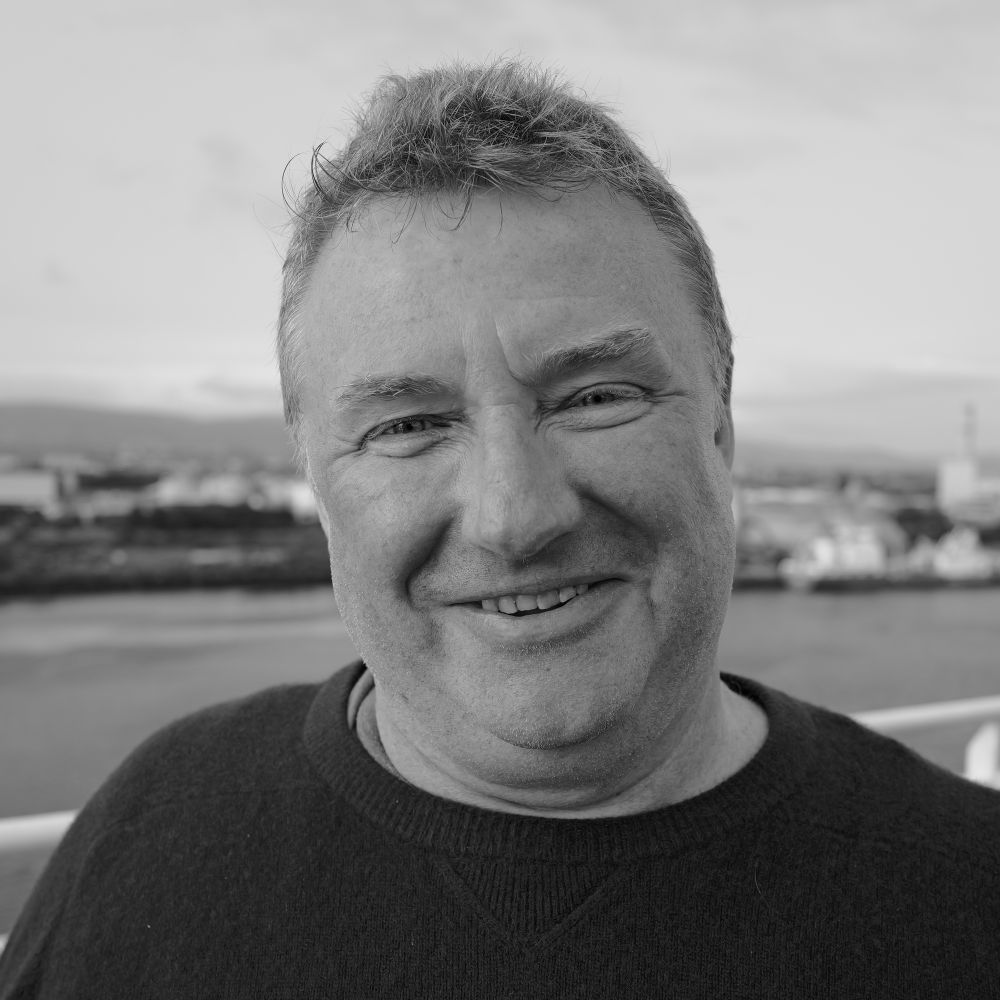 A smiling man wearing a dark sweater stands outdoors near a body of water, with a cityscape and cloudy sky in the background. The image is in black and white.