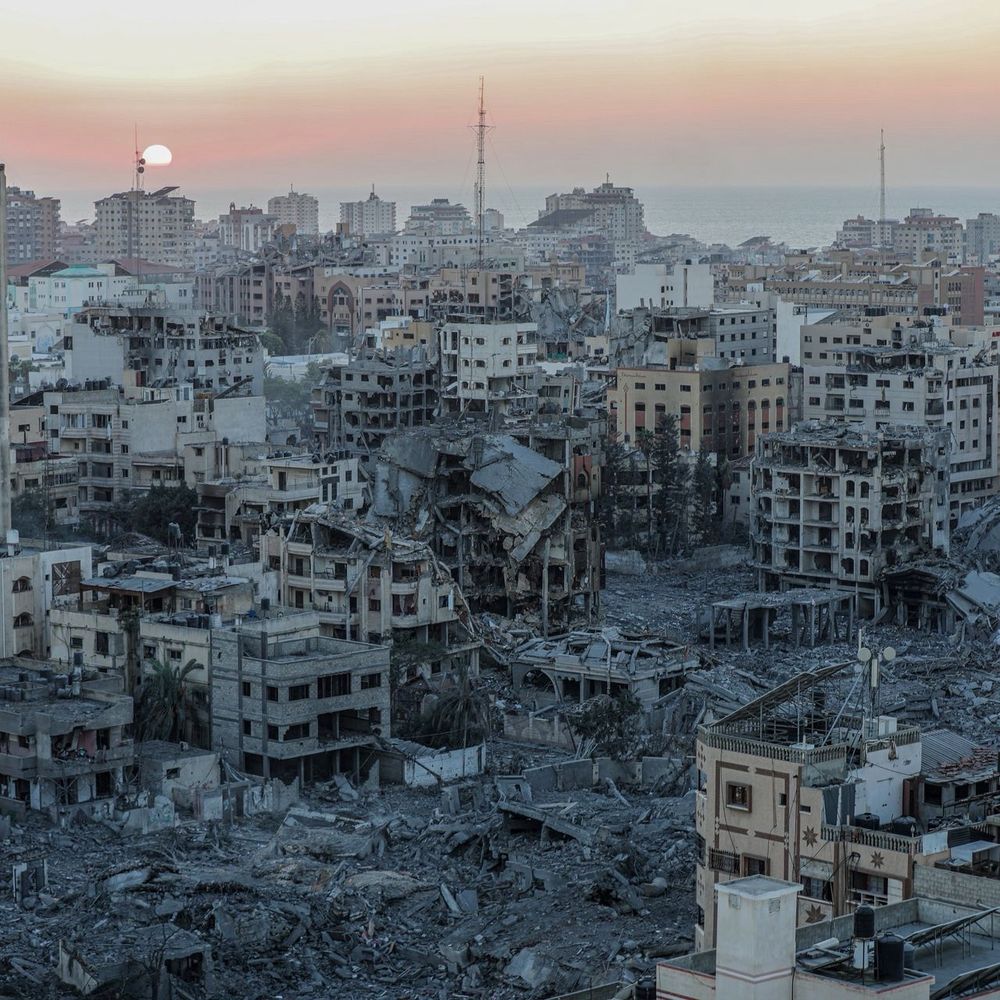 Sunset over a city in Gaza with extensive building destruction and rubble in the foreground, with intact buildings in the background.