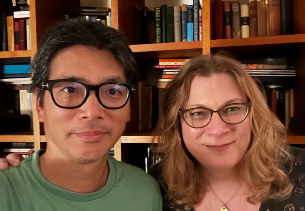 A man and a woman are smiling together in front of a bookshelf filled with books. The man has short black hair and glasses, wearing a green shirt. The woman has wavy, shoulder-length blonde hair and glasses, wearing a dark top.