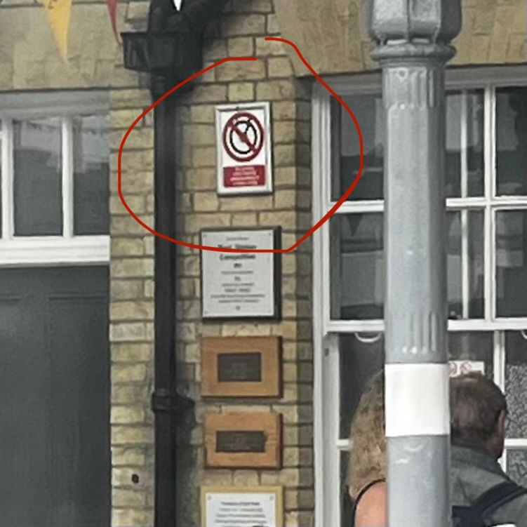 A brick wall features a prominent no something sign, positioned above several plaques and next to a lamppost. There are people partially visible in the foreground.