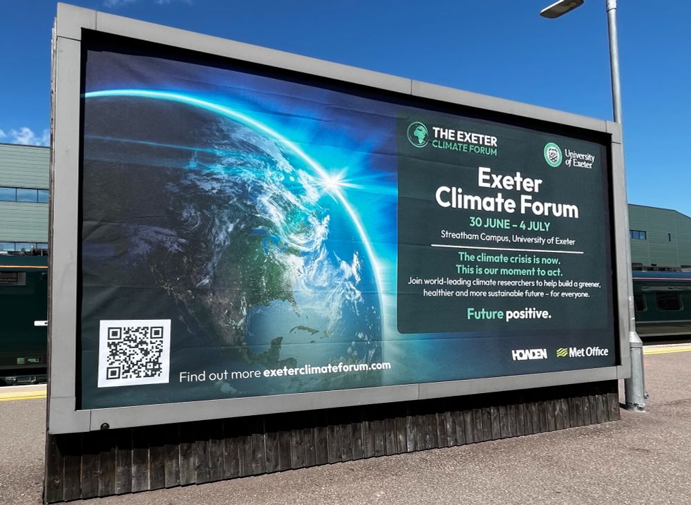 A large billboard advertisement for the "Exeter Climate Forum" is displayed at a train station platform under a clear blue sky. The billboard features an image of Earth from space on the left, showing a vibrant view of North and Central America with a bright sunlight effect on the planet’s horizon. On the right side of the billboard, the text reads:

THE EXETER CLIMATE FORUM
Exeter Climate Forum
30 JUNE – 4 JULY
Streatham Campus, University of Exeter

The climate crisis is now.
This is our moment to act.
Join world-leading climate researchers to help build a greener, healthier and more sustainable future – for everyone.

Future positive.

Below this text, the University of Exeter, HOKDEN, and Met Office logos are shown. A QR code appears in the bottom-left corner of the billboard, with the text:
“Find out more exeterclimateforum.com” next to it. The billboard frame is made of metal and mounted on a wooden base. A green passenger train and a teal train station building are visible in the background.