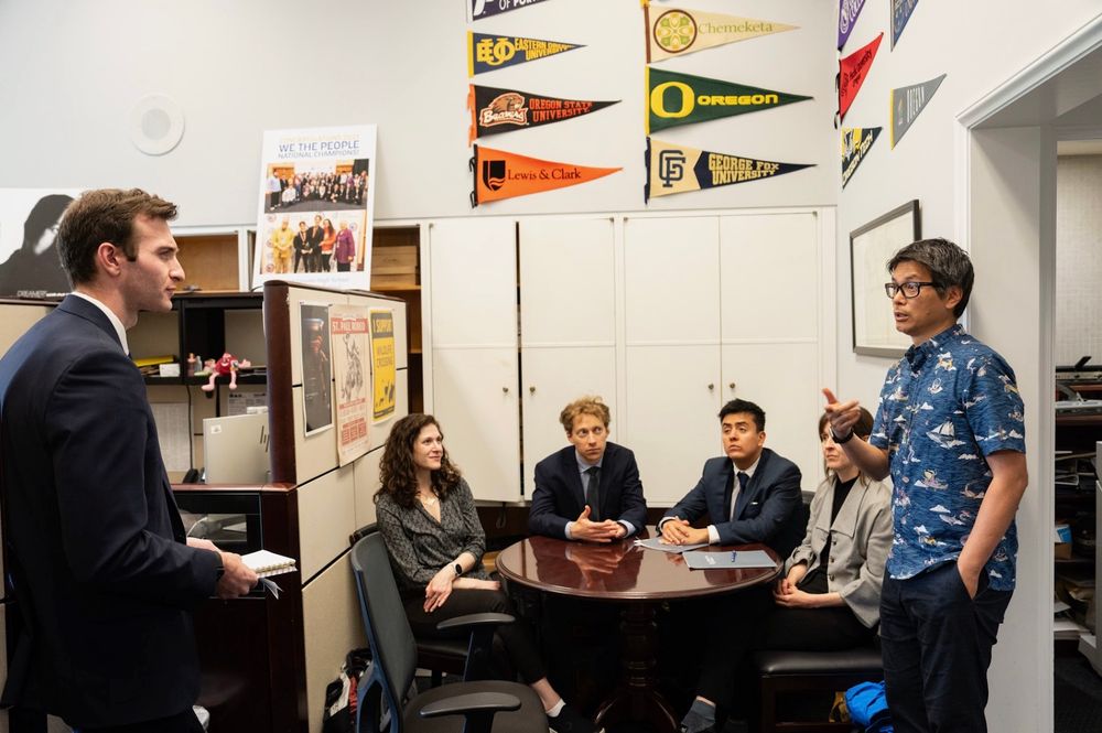 A group of six people is engaged in a discussion in a small office. One person, dressed in a suit, stands and listens, while another in an aloha shirt is explaining something. The others sit around a table, listening attentively. The walls are adorned with various university pennants.