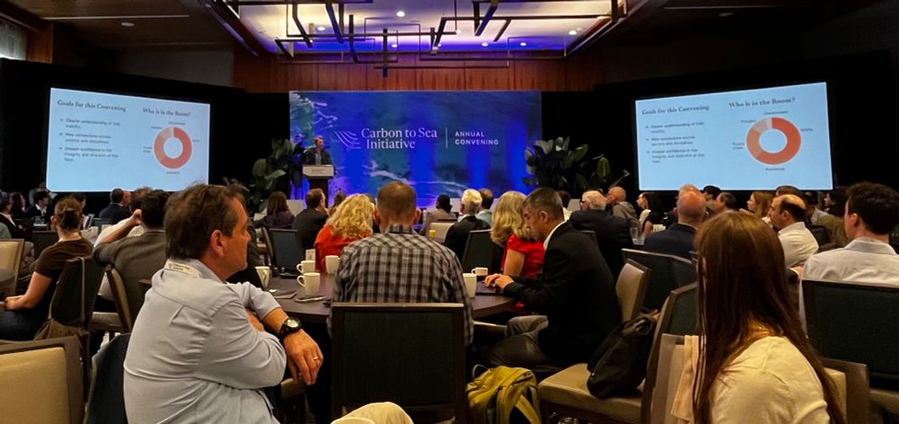 A conference in a modern venue, with attendees seated at round tables. A presenter stands at a podium in front of large screens displaying slides.