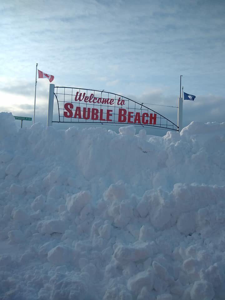 Large red sign spelling Welcome to Sauble Beach (in Ontario, Canada), covered under meters of piled up snow. The sky is blue. 
