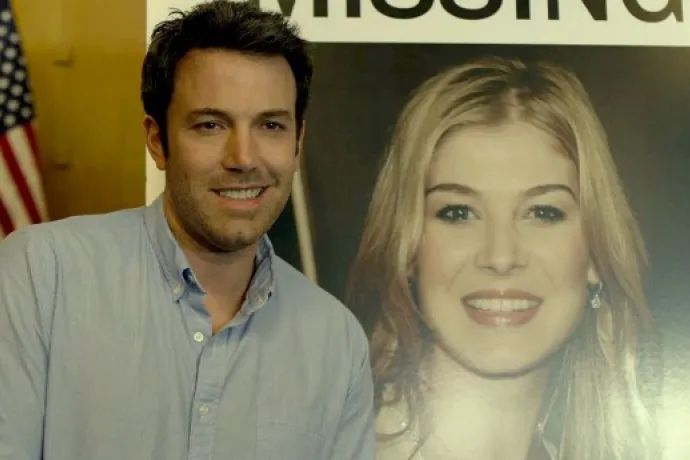 Nick gone girl smiling joylessly next to a large blown up picture of his wife that everyone thinks he’s killed
