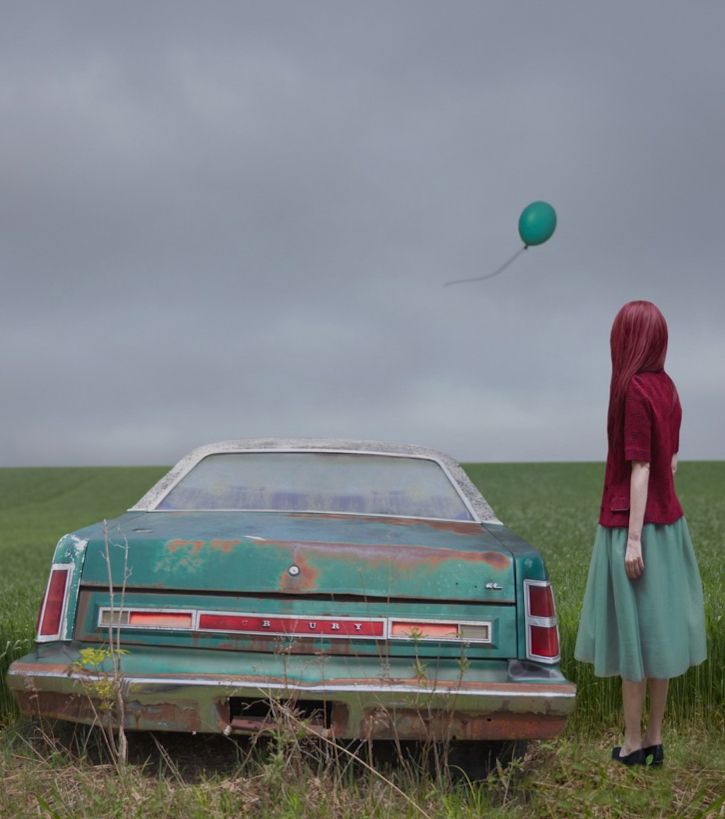 red haired woman standing next to an old turquoise blue mercury sedan in a vast green field
turquoise balloon floats away in the grey sky