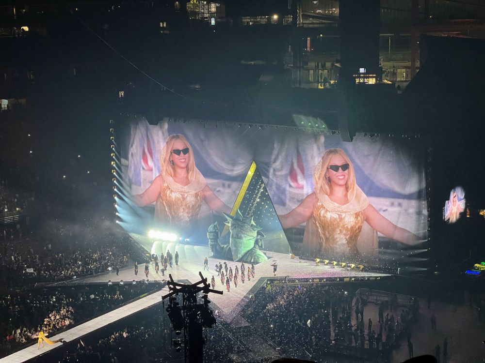 Beyoncé smiling in front of a giant Statue of Liberty wearing a bandana as a mask