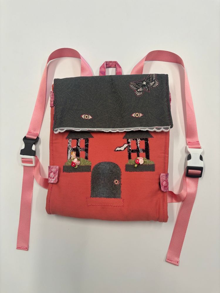 Pink square backpack shaped like a house. The roof, door, and windows are grey and black appliqués. The windows have plant boxes with pink flowers. A girl peels out of one of the windows and there is a bat next to the other window. The roof has a large moth and two eyes. The pink straps have mismatched black and white buckles. 