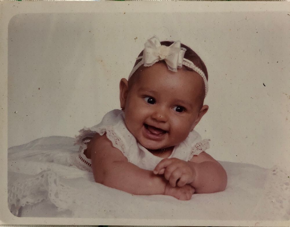 Baby photo of a young baby smiling and resting on a blanket. She is chubby and has dark brown eyes and light brown skin and is wearing all white, including a white bow. 