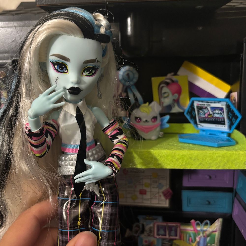 photo of a Frankie stein doll posed with their hand to their face. They have light blue skin, white and black hair with half loose and half in a Dutch braid, heavy pink and yellow eye makeup, & black lipstick. They are wearing a white collared sleeveless top with pink and blue trim, pink sleeves with black white and lime lightning stripes, and pleather black pants with pink blue and yellow accents. The background is of a doll room with a nonbinary flag and a poster of Frankie’s with their blue laptop and a plush of their dog. 