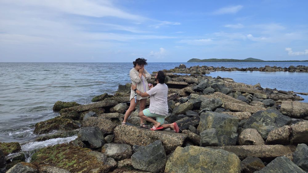Photo of a proposal on a rocky beach; the person being proposed to has their hands over their mouth in surprise. The person proposing has brown skin and curly dark hair in a ponytail; he’s wearing a cream linen shirt, mint swim trunks, and red adidas slides. The person being proposed to has light brown skin and curly dark hair with bangs in a ponytail; they are wearing a beige linen beach cover up and rainbow adventure sandals. 