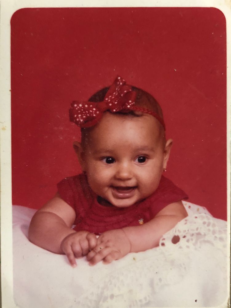 Baby photo of a young baby smiling and resting on a blanket. She is chubby and has dark brown eyes and light brown skin and is wearing all red, including a red bow. 