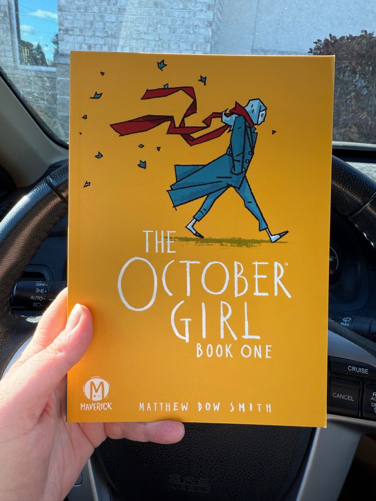 A photograph of the newly released graphic novel The October Girl, by Matthew Dow Smith. The cover is primarily a golden yellow, with an illustration of a young woman walking in the autumn wind.