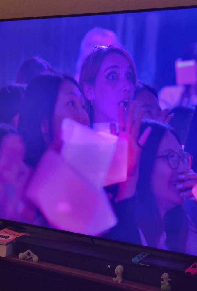 A tall woman with a shocked look on her face in a crowd of people holding a square light stick. 