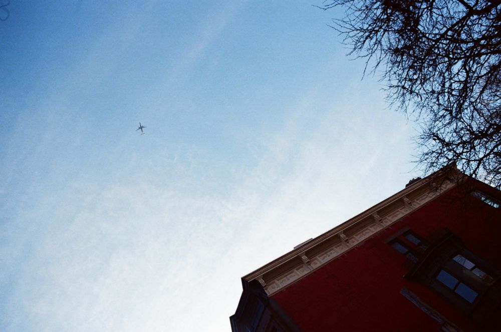 A plane tiny and high in the blue sky turning yellow at dusk above a red brick building and leafless tree.