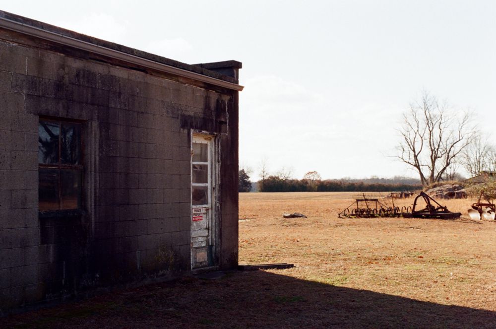 A dilapidated brick shed with a faded white door in shadow casts a shade across brown grass, rusted metal from farm equipment in the background.