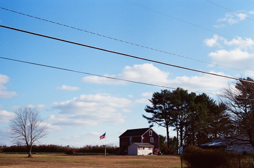 A white shed in front of a red barn near tall trees in a brown grass field, an American flag lifted in the wind.