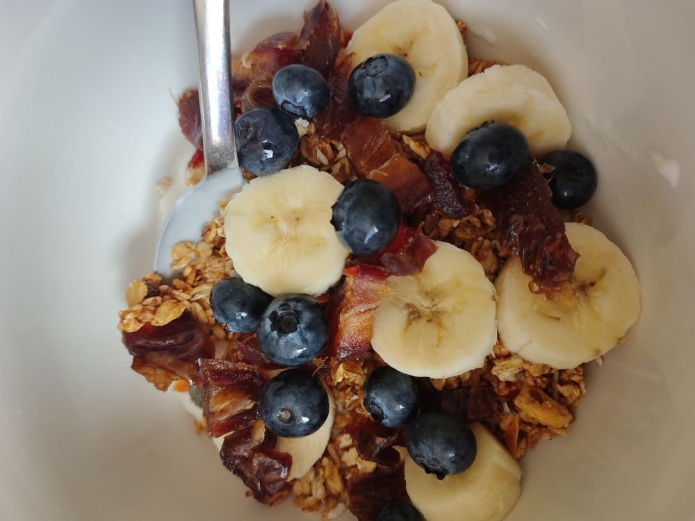 A white bowl with a spoon at 11 o'clock. A mix of blueberries, sliced banana and dates on top of light brown granola with dairy free coconut yoghurt beneath.