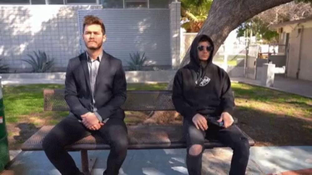 a man in a suit sits next to a man in a black hoodie with the word raiders on it