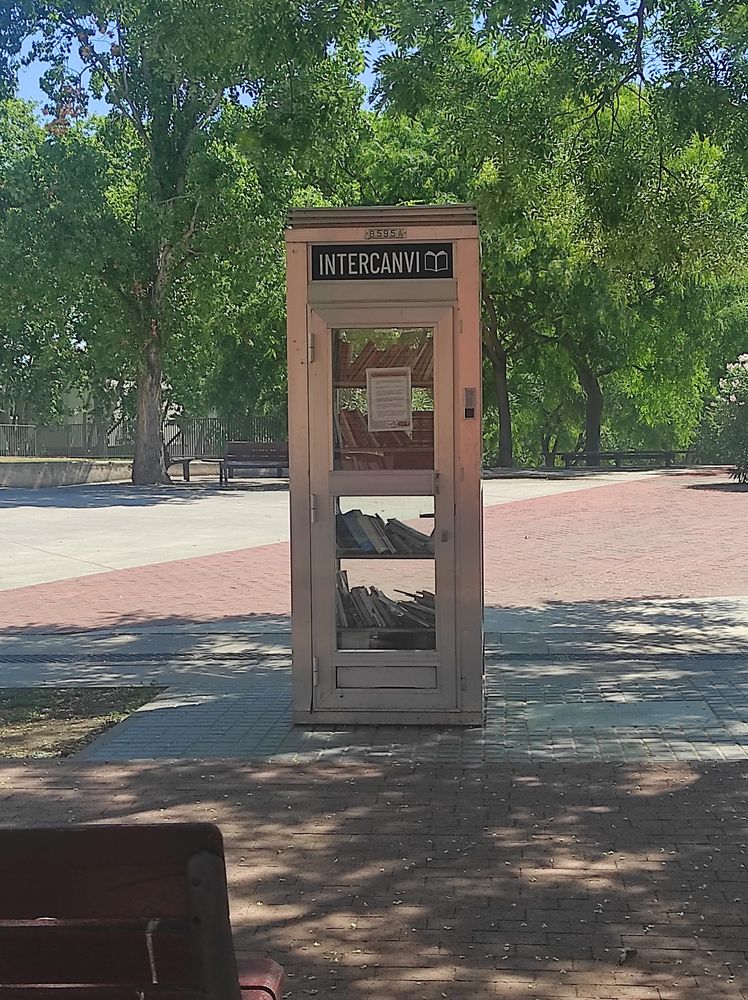 Foto de una antigua cabina telefónica de aluminio y cristal con estanterías en el interior llenas de libros, en una plaza pública. En la parte superior de la cabina, sobre la puerta de entrada, hay un letrero que pone Intercanvi y la silueta de un libro abierto. 
Unos frondosos árboles dan sombra a la antigua cabina y decoran los adoquines del suelo con pequeñas flores blancas. 