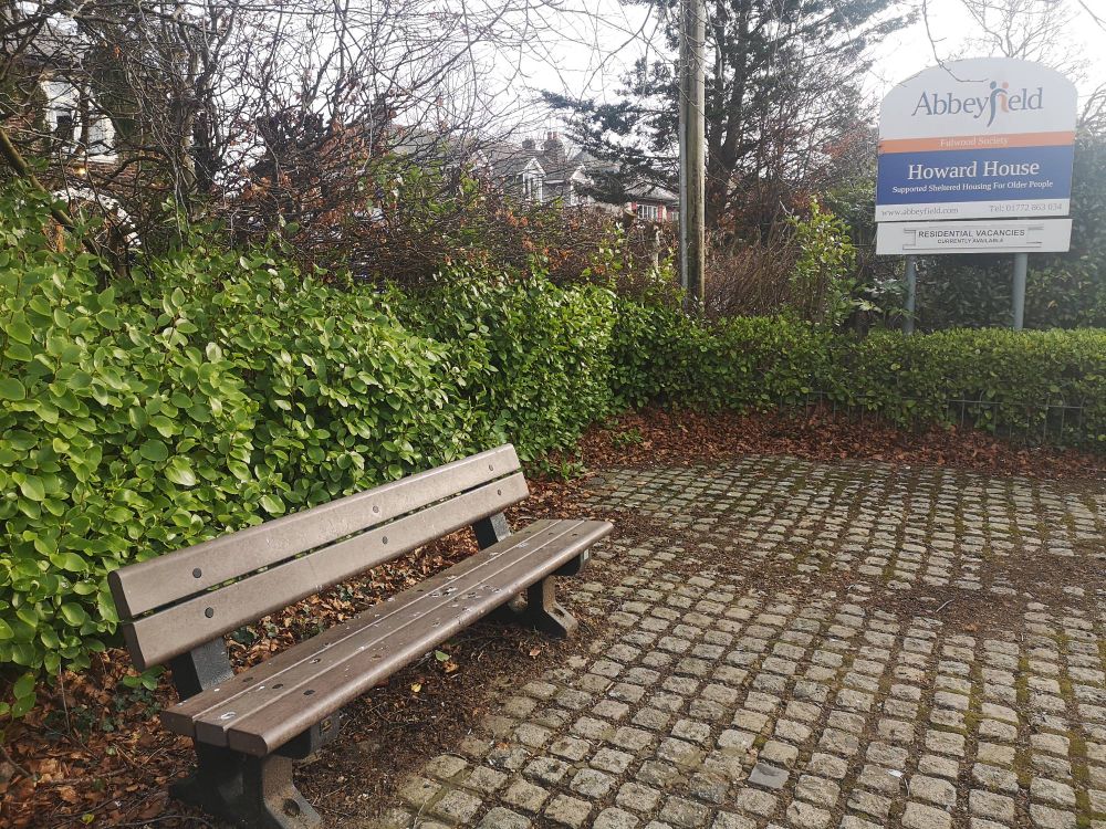 Bench with bird poo on it next to Howard House residential home on Garstang Road. The bench is set on a cobbled area and surrounded by shrubbery.