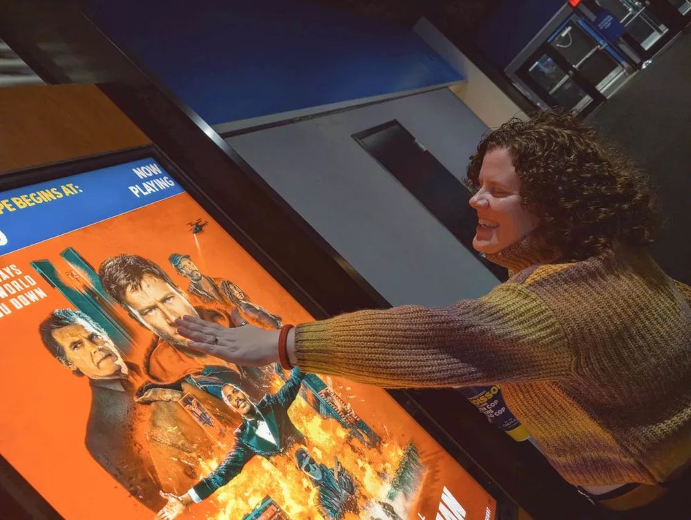 me, megan aka tat, a woman with short, curly brown hair is standing in a movie theatre hallway, smiling with their eyes closed as they lean forward to place a hand on a brightly lit movie poster display for the running man. they’re wearing a chunky, multicolored knit sweater in shades of mustard yellow, brown, and tan. the poster they’re touching is dominated by vivid orange tones and features dramatic action imagery: several men in intense poses, explosions, and helicopters. megan's outstretched arm and hand are pressed gently over the main character’s (glen powell) face on the poster, as if playfully or affectionately reaching for him. the theatre surroundings are dim, with dark walls, closed auditorium doors, and overhead lighting casting a warm glow on the person’s face.