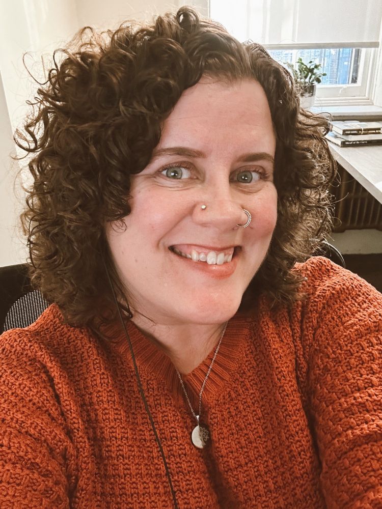Megan with short, curly brown hair smiles at the camera in a warmly lit indoor setting. They’re wearing a textured rust-colored sweater, a silver necklace with round pendants, and have both a nose stud and hoop. Behind them, there’s a window letting in daylight, a small green plant, and a desk with a few stacked books.