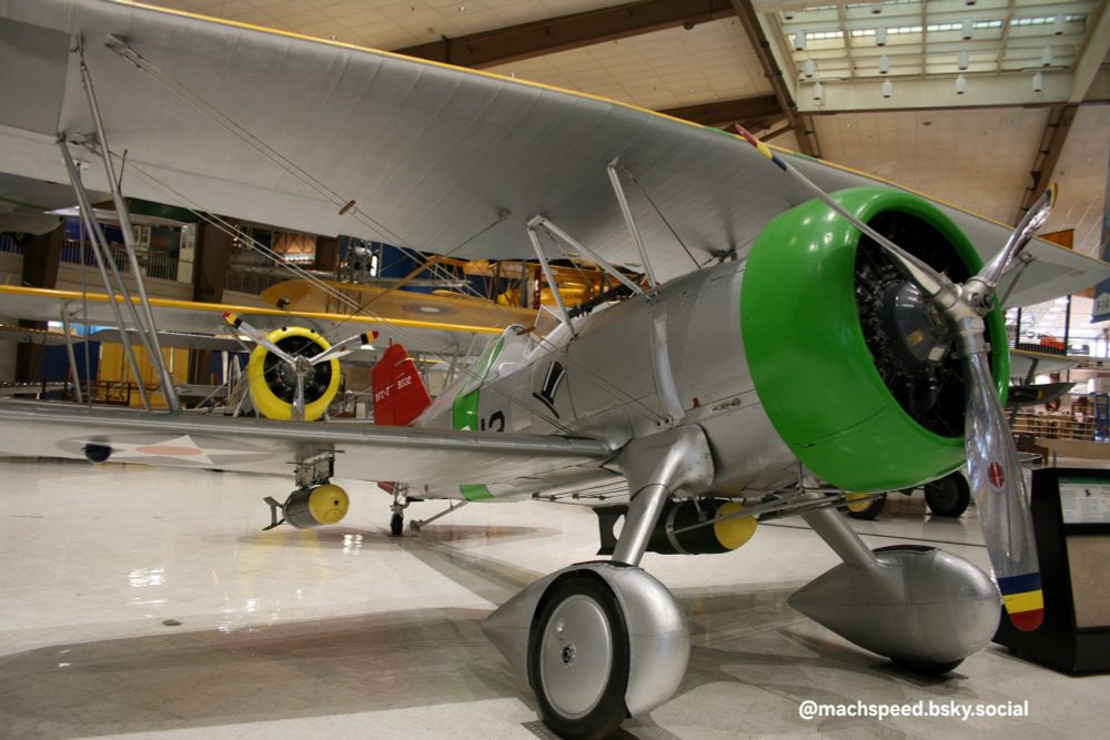 BFC-2 Goshawk, early US divebomber. The arm that swings the centerline ordnance clear of the prop in a dive is visible.
A silver bi-plane with bright green engine cowling, vertical green stripe behind the cockpit, and red tail fin. A dashing black and white top hat squadron patch is painted on the side of the plane. 
Photo by @machspeed.bsky.social