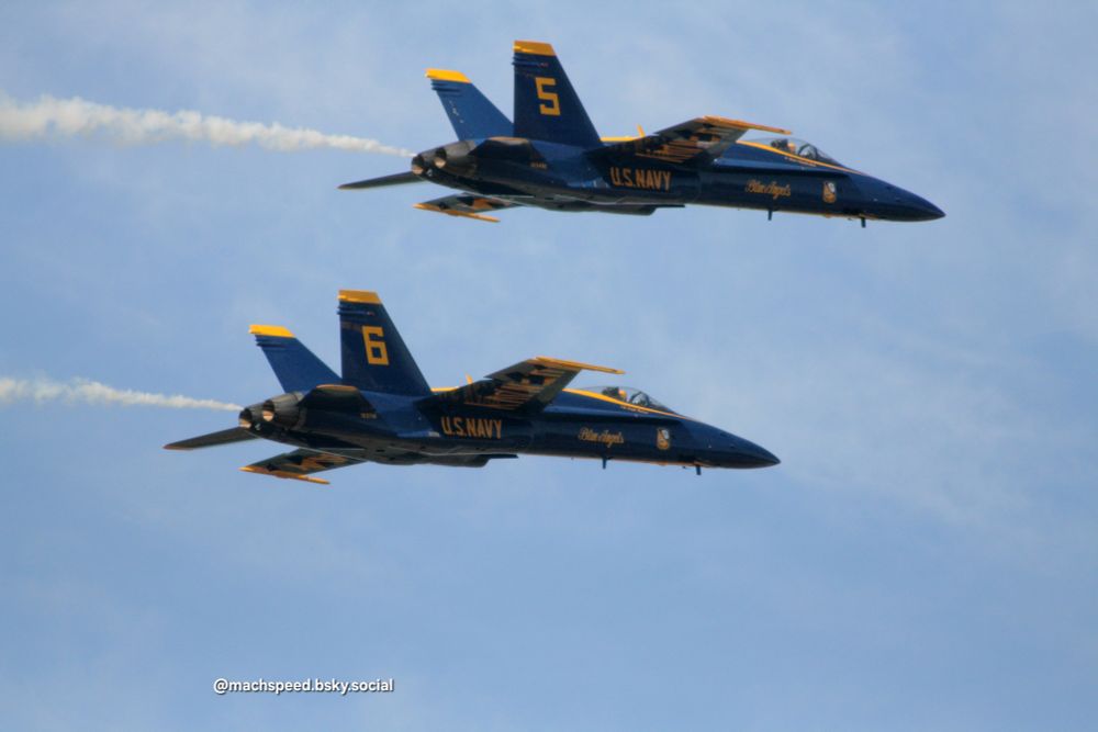 Two Blue Angels streak past in F/A-18C Hornets. Brilliant blue with golden yellow trim, the Blues always look nice in a sunny sky. One sports a large yellow #5 on the tail, the other a #6. Photo 2016 by @machspeed.bsky.social
