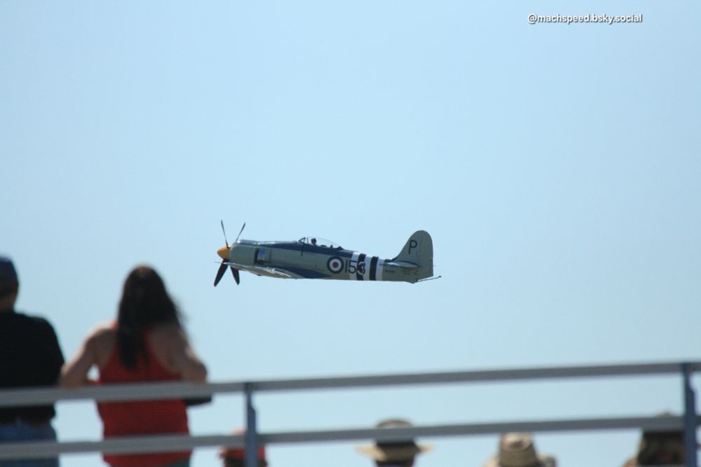 Hawker Sea Fury FB Mk. 11 flying past with bleachers and spectators out of focus in the foreground. There is a Blue, white, and red RAF roundel on the the fuselage with the number 153 over vertical black and white invasion stripes. Prop blur will cost you extra. 
Eglin AFB 75th Anniversary airshow April 11, 2010. Photo by @machspeed.bsky.social