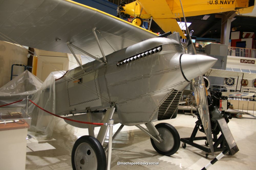 Curtis F6C-1 Hawk biplane in shiny silver livery with yellow on top of upper wing. From the 1 o'clock postion just right of the nose. Undergoing restoration at the time, the rear of the plane is covered in a clear tarp. A beautiful airplane. Nat. Naval Aviation Museum NAS Pensacola. Photo by @machspeed.bsky.social