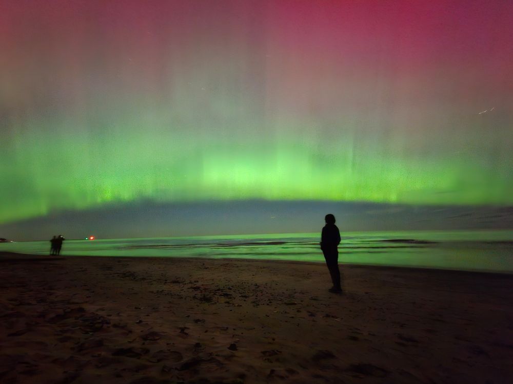 a photograph of the northern lights over lake ontario. A figure stands on the beach facing away from the camera, looking at the sky. She is thinking about yaoi