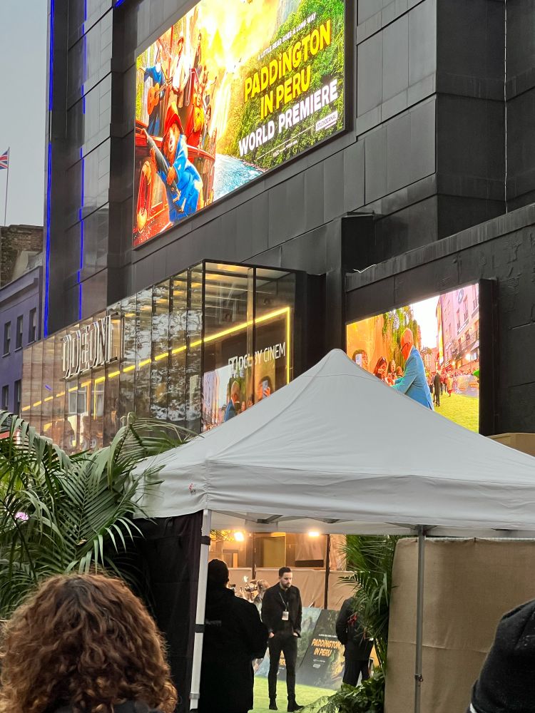 A picture of a tent and barriers outside the world premiere of the Paddington in Peru movie, with a peek of the green carpet and the actors shown on a monitor that can be seen from outside the barriers 
