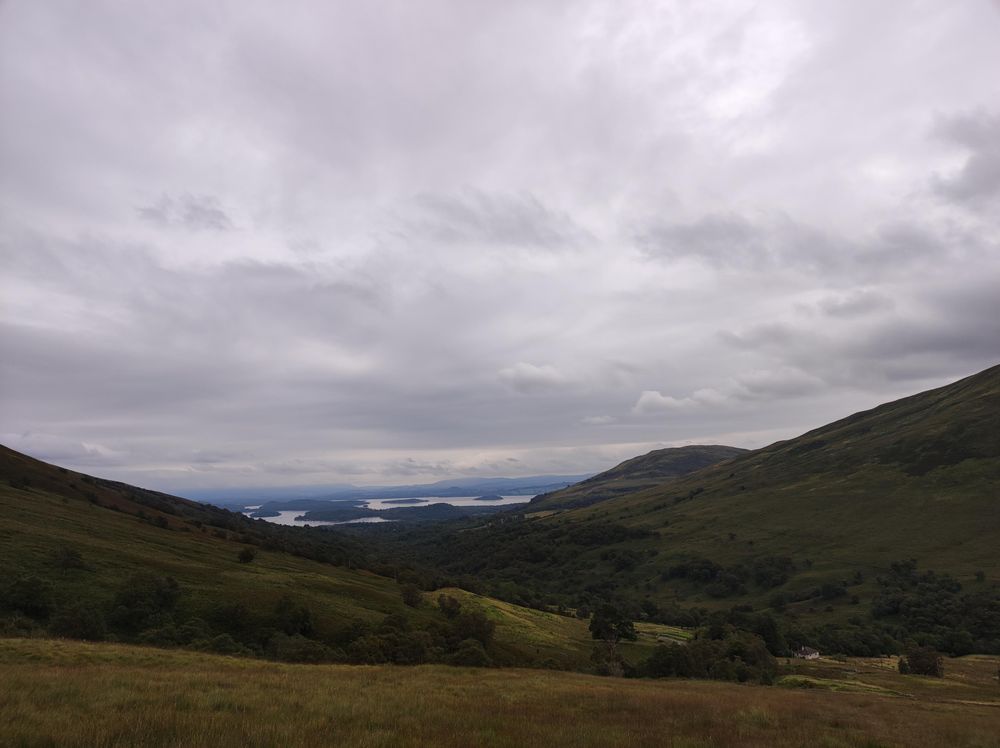 Photo of green hills from a somewhat elevated position. In the background you can see a lake. The sky is covered in clouds.
