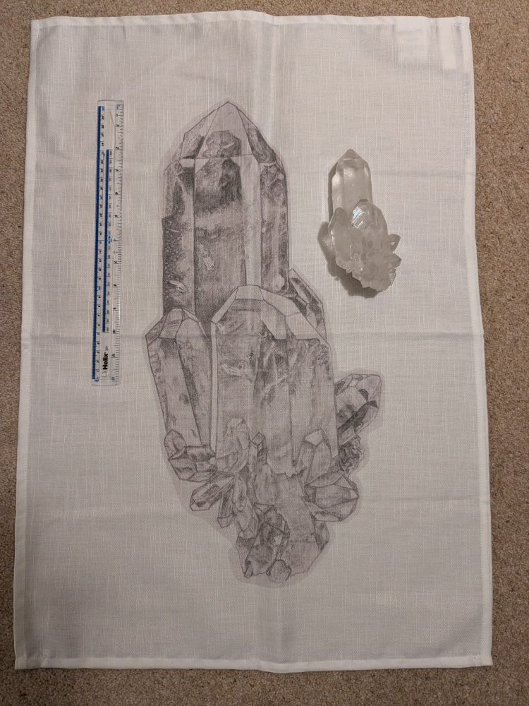 Photograph of a large tea towel with a fairly loose-looking weave. There is a large pencil drawing of a crystal in the centre of the tea towel. To the left is a 30 cm ruler for scale, showing that the length of the drawing is around 50cm. To the right is a cluster of quartz crystals about 15 cm long.