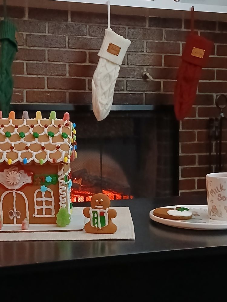 A decorated gingerbread house, gingerbread man along with a glass of milk and a cookie left for Santa in front of a fireplace with stocking hung from the mantle.