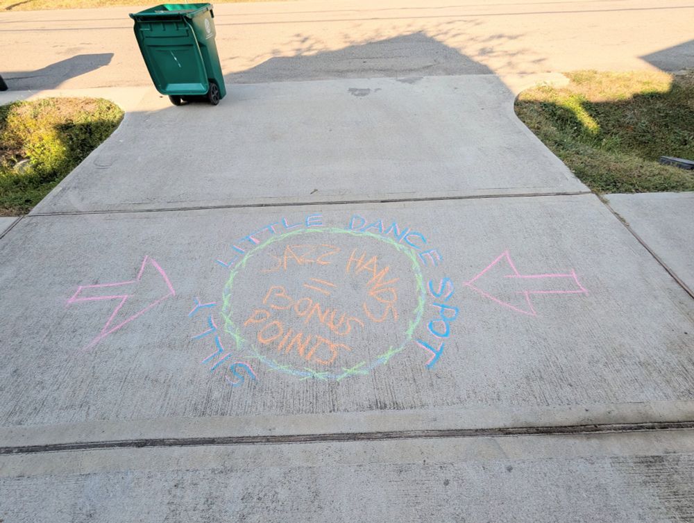 A chalk circle on a driveway that reads "Silly Little Dance Spot - Jazz Hands= Bonus Points" and two pink arrows pointing to it