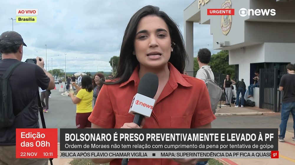 Plantão da globonews na porta da prisão do bolsonaro. Atrás da repórter, uma senhora de blusa amarela toma champagne em uma taça