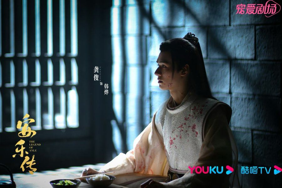 Gong Jun stars as Han Ye in Legend of Anle. He sits in shadow in a jail cell wearing white robes that have been splattered with blood drops that resemble red flowers.
