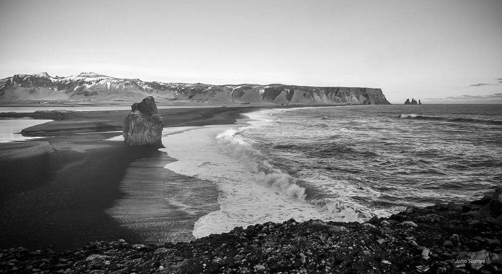 Waves rolling on to a black sand beach with a large rock formation (Arnardrangur) rising out of the sand. There is rocks in the foreground and a long peninsula with patches of snow along the back of the image. Taken on the south shore of Iceland in February.