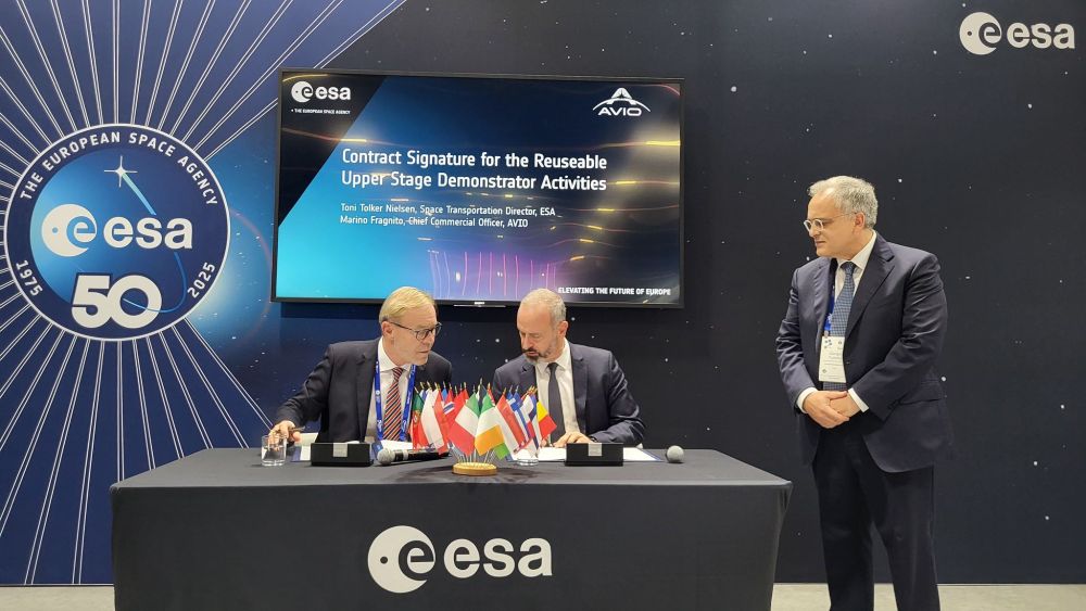 A ceremonial contract signing event is taking place, highlighting a collaboration on the "Reusable Upper Stage Demonstrator Activities." Two key representatives: ESA's Director of Space Transportation Toni Tolker-Nielsen (left) and Avio’s Chief Commercial Officer and Launch Services Director Marino Fragnito (right), both seated at a table, signing the agreement. The table is decorated with miniature flags symbolising international cooperation. ESA's Chief Technical Advisor for Space Transportation Giorgio Tumino stands nearby, observing the proceedings. Behind them, a large screen displays the event title and participant names, with ESA and AVIO logos, along with a commemorative emblem marking ESA’s 50th anniversary in 2025. The setting conveys a formal and significant milestone in space transportation development.