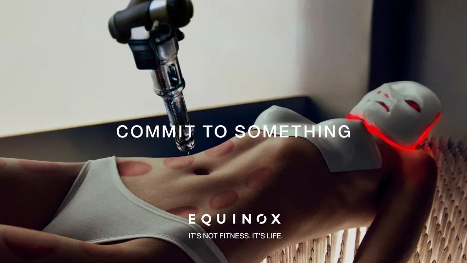 This image appears as it does in an actual Equinox ad, with the text COMMIT TO SOMETHING overlaid. I do not know what we are actually looking at, but it appears to be a cis woman enduring numerous beauty treatments and plastic surgeries simultaneously while on an illuminated bed of nails.