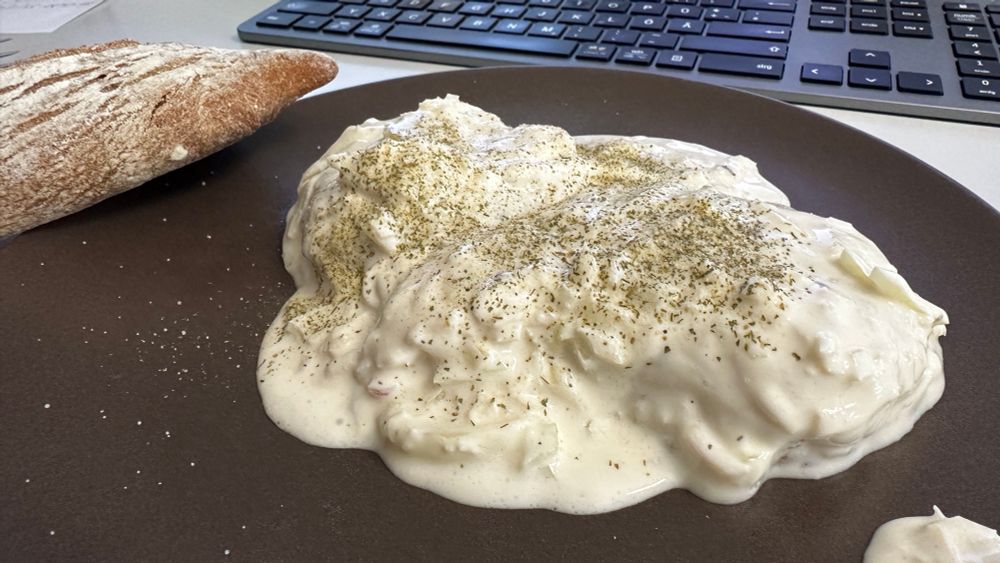 Brauner Teller, unter schmand und Zwiebeln versteckter handkäs, daneben liegt ein Brötchen, alles auf einem Schreibtisch, die Tastatur ist im Hintergrund zu sehen.