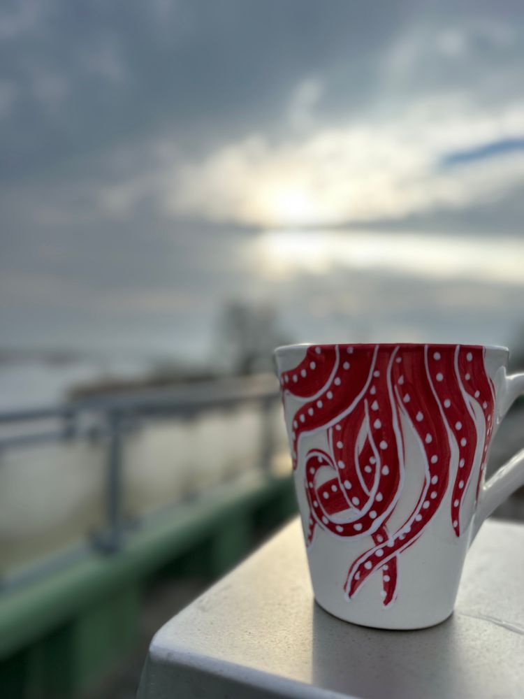 Weisse Tasse mit roten Tentakeln, dahinter verschwommen der Sonnenaufgang.