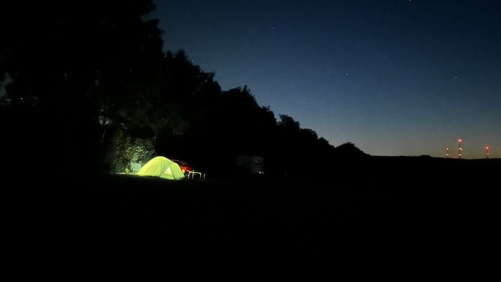 Nachtaufnahme einer Wiese auf der ein Zelt steht, grün, in dem Licht brennt und damit das Zelt zum strahlen bringt.